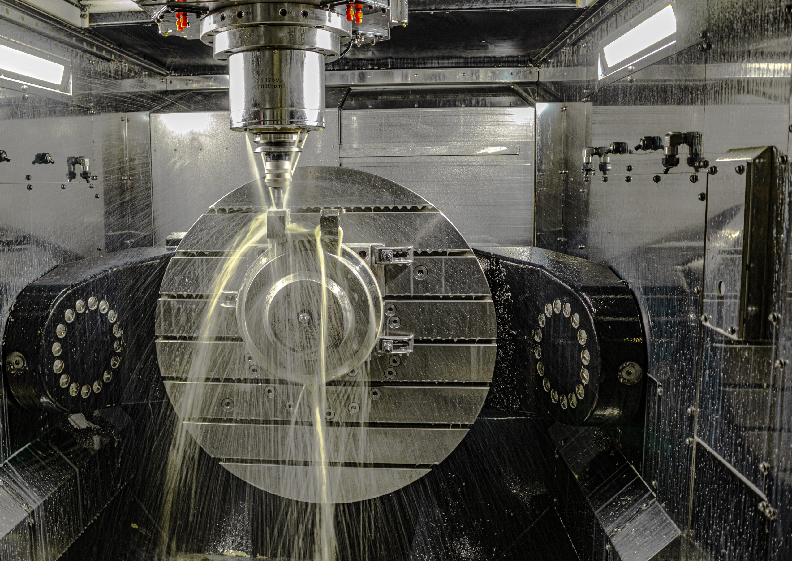 A large milling machine processes a metal part. The photo was taken while the machine was running. The oil jet cools the cutter and the part.